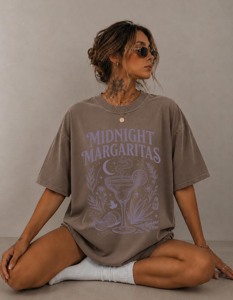 K&ouml;nnte beinhalten: Ein braunes Oversize-T-Shirt mit dem Schriftzug Midnight Margaritas in hellvioletter Schrift. Die Grafik zeigt ein zentrales Margaritaglas, eine Mondsichel, Zitrusfr&uuml;chte und botanische Elemente in einem passenden hellvioletten Farbton.