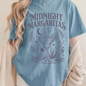 K&ouml;nnte beinhalten: Ein blaues Kurzarm-T-Shirt mit dem Text MIDNIGHT MARGARITAS in dunkelvioletter Schrift. Die Grafik zeigt ein Margaritaglas mit Dampf, eine Mondsichel, Sterne, Limettenscheiben, eine Agavenpflanze und verschiedene kleine Blumenmotive.