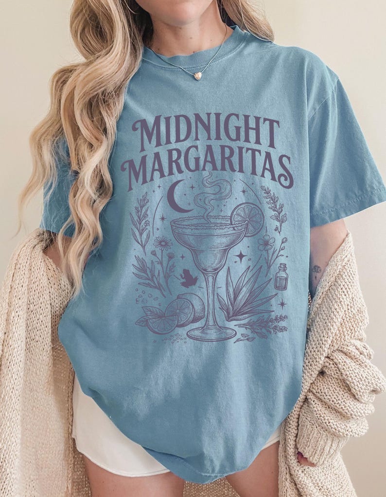 K&ouml;nnte beinhalten: Ein blaues Kurzarm-T-Shirt mit dem Text MIDNIGHT MARGARITAS in dunkelvioletter Schrift. Die Grafik zeigt ein Margaritaglas mit Dampf, eine Mondsichel, Sterne, Limettenscheiben, eine Agavenpflanze und verschiedene kleine Blumenmotive.