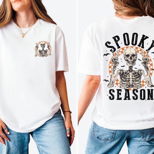 Puede incluir: Camiseta blanca con un gr&aacute;fico de esqueleto. La parte delantera presenta un peque&ntilde;o dise&ntilde;o de esqueleto, mientras que la parte trasera muestra un esqueleto m&aacute;s grande con las palabras "Spooky Season" en negro, con una fuente desgastada. El dise&ntilde;o incluye murci&eacute;lagos y un c&iacute;rculo naranja a cuadros.