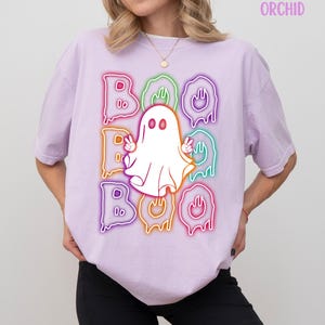 Pode incluir: Camiseta lilás claro com um design gráfico com a palavra "BOO" em letras de néon coloridas e um fantasma branco fazendo o sinal de paz. A palavra "ORCHID" é impressa no canto superior direito.