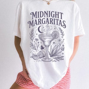 K&ouml;nnte beinhalten: Ein wei&szlig;es Kurzarm-T-Shirt mit einem dunkelvioletten Grafikdruck mit dem Text "MIDNIGHT MARGARITAS" &uuml;ber einer Illustration eines Margaritaglases, einer Mondsichel, Sternen, Limettenscheiben, einer Agavenpflanze und einem kleinen Frosch.