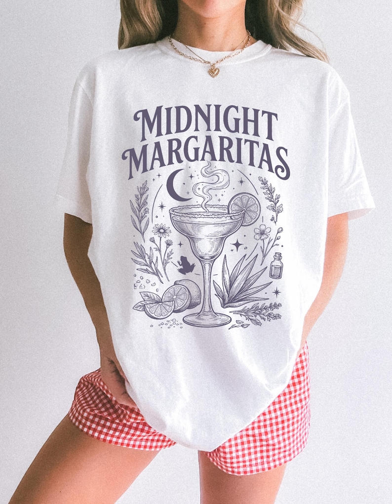 K&ouml;nnte beinhalten: Ein wei&szlig;es Kurzarm-T-Shirt mit einem dunkelvioletten Grafikdruck mit dem Text "MIDNIGHT MARGARITAS" &uuml;ber einer Illustration eines Margaritaglases, einer Mondsichel, Sternen, Limettenscheiben, einer Agavenpflanze und einem kleinen Frosch.