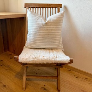 Beige-grau gestreifte Kissenhülle auf einem Holzstuhl mit heller Sitzauflage in einer schlichten Wohnecke.