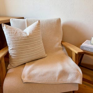 Beige-grau gestreifte Kissenhülle auf einem hellen Holzsessel mit cremefarbener Strickdecke in einer warmen, neutralen Wohnecke.