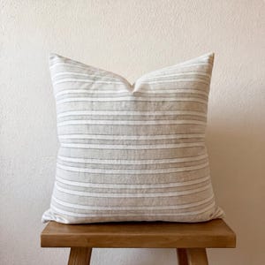 Beige-grau gestreifte Kissenhülle 50 × 50 cm, frontal auf einem Holzstuhl vor heller Wand. Die Hülle ist für das Foto mit einem Füllkissen dekoriert.