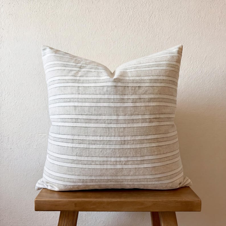 Beige-grau gestreifte Kissenhülle 50 × 50 cm, frontal auf einem Holzstuhl vor heller Wand. Die Hülle ist für das Foto mit einem Füllkissen dekoriert.