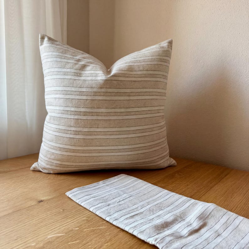 Beige-grau gestreifte Kissenhülle auf einem Holztisch mit gefaltetem Stoffmuster im Vordergrund; Webstruktur und Farben sind gut erkennbar.
