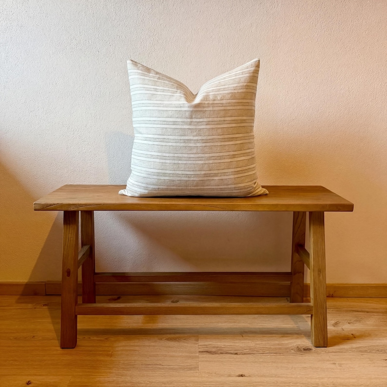 Gefüllte Kissenhülle in Beige und Grau auf einer schlichten Holzbank vor heller Wand; Vorderseite, quadratische Form und Größenwirkung sind sichtbar.