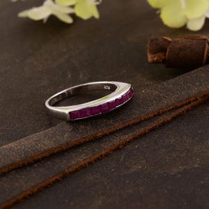 May include: A silver ring featuring a line of rectangular, deep red gemstones. The ring is set on a dark brown leather surface, with a leather strap and some white and yellow flowers in the background.