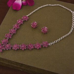 Puede incluir: Un conjunto de collar y pendientes a juego de color plateado. La joyería presenta un diseño floral con racimos de pequeñas piedras preciosas de color rosa intenso. El collar está colocado sobre una superficie marrón, con una flor rosa en el fondo.