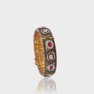 May include: A gold and silver hinged bracelet with alternating ruby and diamond-like stones. The bracelet has a detailed filigree design on the inside and a secure clasp. The stones are set in a dark metal, creating a striking contrast.