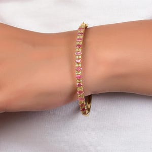May include: Gold-toned bracelet with alternating pink and yellow rectangular gemstones. The bracelet is worn on a wrist against a white background. The gemstones are set closely together, creating a continuous line of color.