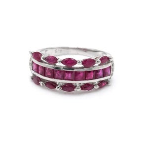 May include: A silver ring with three rows of vibrant red gemstones. The center row features rectangular stones, while the top and bottom rows have oval-shaped stones. The ring is set against a white background, highlighting the jewelry's design.
