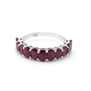May include: A silver ring with a line of oval-cut, deep red gemstones. The gemstones are set close together, forming a continuous band of colour. The ring is shown against a white backdrop.