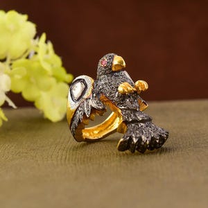 Peut inclure: Bague dorée ornée d'un oiseau stylisé. Le corps de l'oiseau est noir avec des détails dorés, un œil en pierre rouge et une pierre blanche sur le corps de la bague. Fond flou.