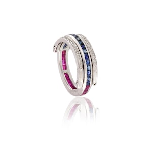 May include: A silver ring with three bands. One band is lined with small, square-cut rubies, another with blue sapphires, and the third with small diamonds. The ring is set against a white background.