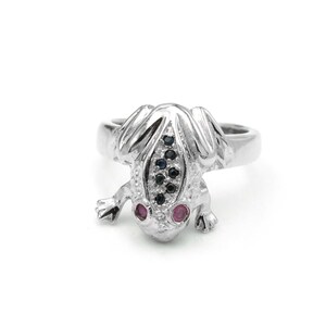 May include: A silver-toned frog ring with a textured surface. The frog's eyes are pink gemstones, and the back has a row of black and blue gemstones. The ring is set against a white background.