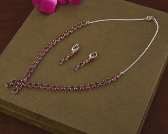 Puede incluir: Un conjunto de collar y pendientes de plata con piedras preciosas rojas oscuras de corte ovalado. El collar y los pendientes tienen un diseño a juego. El conjunto de joyas se presenta sobre una superficie marrón, con una flor rosa en el fondo.