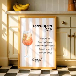 Op de afbeelding: Een ingelijste wandkunstprint met een achtergrond van verticale witte en perzikkleurige strepen. Het toont een illustratie van een oranje cocktail in een glas op voet en zwarte tekst met Aperol spritz BAR, receptinstructies en Enjoy!.
