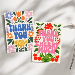 Op de afbeelding: Twee wenskaarten met bloemmotieven. Eén kaart zegt "THANK YOU" in blauwe bubbelletters omringd door oranje, paarse en roze bloemen. De andere kaart zegt "THANK YOU CHERRY MUCH" in roze bubbelletters, met kersen en rode bessen.