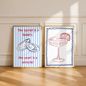 Op de afbeelding: Twee ingelijste kunstdrukken. Eén heeft een blauw-wit gestreepte achtergrond met een oesterillustratie en de tekst "The oyster is a luxury, the pearl is a miracle!". De andere print toont een cocktailglas met een schijfje citroen.
