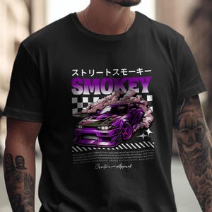 May include: Black t-shirt featuring a purple sports car with smoke billowing from the tires. The design includes Japanese text and the word "SMOKEY" in a gradient purple color. The shirt also has additional text at the bottom.