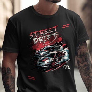 以下が含まれることがあります： 黒いTシャツ。ドリフトする2台の車のグラフィックと「STREET DRIFT」の文字。赤と白のアクセント、煙の効果、スピードラインが含まれており、レースをテーマにしています。