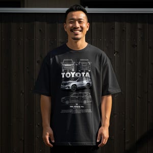 Può includere: T-shirt nera con il logo Toyota e illustrazioni di veicoli Toyota. È visibile anche il testo "THE TOYOTA TXL". La persona nella foto sta sorridendo.
