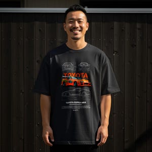 May include: Black t-shirt featuring a graphic of a Toyota Supra MK4 car in orange and white. The shirt also displays the Toyota logo and car blueprints. The model is wearing the shirt.