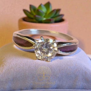 May include: A silver engagement ring with a large, clear, round-cut diamond. The ring has dark purple accents on the band. It's displayed on a light purple cushion, with a small potted succulent in the background.