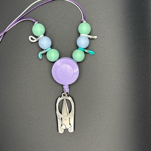 Puede incluir: Un cordón con cuentas con un carrete retráctil morado y un dije de tijeras plateadas. El cordón presenta cuentas verdes y celeste, con cordones blancos y morados. El dije de tijeras es detallado y tiene un diseño único.