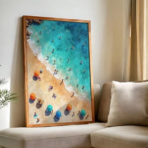 May include: A framed art print depicts a vibrant beach scene. Turquoise water meets a sandy shore, with colorful umbrellas and figures scattered across the beach. The artwork is encased in a natural wood frame, adding a touch of warmth to the coastal theme.