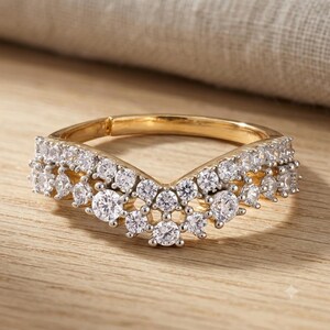 Könnte beinhalten: Ein goldener Ring in Chevron-Form mit zwei Reihen klarer, rund geschliffener Steine entlang der Ringschiene. Das Design bildet ein V, wobei die Steine in einem geschichteten Muster angeordnet sind, das dem Verlauf des Rings folgt, mit metallischem Glanz.