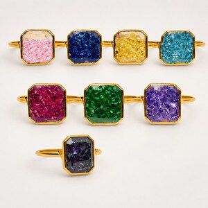 May include: A collection of seven gold-toned rings, each featuring a rectangular setting filled with textured, crystalline material in various vibrant colors including pink, dark blue, yellow, light blue, magenta, green, purple, and dark violet.