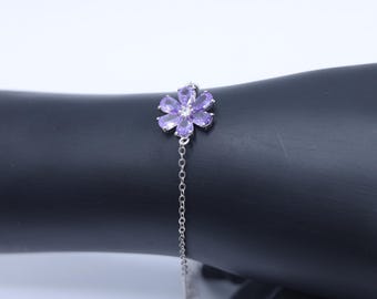 May include: A silver bracelet featuring a flower design with purple gemstones. The flower is the focal point, with a delicate chain extending from it. The bracelet is displayed against a dark background, highlighting its details.
