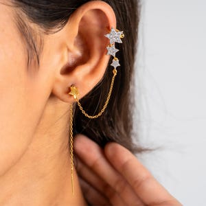 Puede incluir: Juego de pendientes y puño de oreja dorados con diseños de estrellas. El puño de oreja tiene tres estrellas con piedras preciosas transparentes, conectadas por una delicada cadena a un pendiente de estrella. El pendiente tiene una barra larga y colgante.