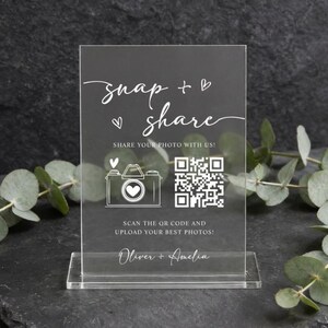 Könnte beinhalten: Ein durchsichtiges, rechteckiges Acrylschild mit weißem Aufdruck, der snap + share, SHARE YOUR PHOTO WITH US!, SCAN THE QR CODE AND UPLOAD YOUR BEST PHOTOS! sowie Oliver + Amelia lautet, ergänzt durch ein Kamerasymbol und einen QR-Code.