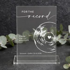 Op de afbeelding: Een rechthoekig bord van helder acryl met witte bedrukte tekst die luidt FOR THE record, Please sign our guestbook, en Savanah + Griffin | 10.10.2026. Een witte afbeelding van een vinylplaat met een klein hartje in het midden is inbegrepen.