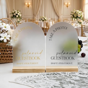 Puede incluir: Dos letreros arqueados con bases de madera. Los letreros dicen "TAKE A PHOTO!" y "polaroid GUESTBOOK SNAP IT. STICK IT. SIGN IT." en letras doradas y negras. El fondo muestra una recepción de boda.