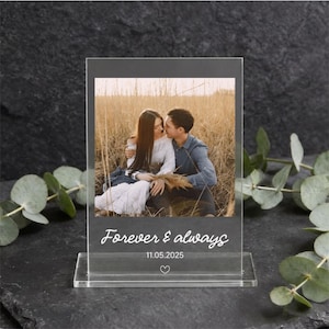 Può includere: Una cornice portafoto rettangolare in acrilico trasparente con una fotografia stampata di una coppia in un campo. Il testo Forever & always è stampato in bianco sotto la foto, seguito dalla data 11.05.2025 e da una piccola icona a forma di cuore.