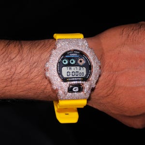 Könnte beinhalten: Eine Casio G-Shock-Uhr mit gelbem Armband und einem Zifferblatt, das mit kleinen, funkelnden Steinen besetzt ist. Die Digitalanzeige zeigt die Uhrzeit und andere Funktionen an. Die Uhr befindet sich an einem Handgelenk.