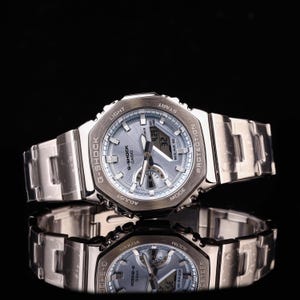 May include: A silver G-Shock Casio watch with a light blue face and a stainless steel strap. The watch displays the time and date. The watch is reflected in the black surface below.