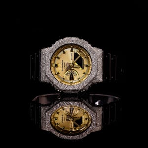 May include: A G-Shock Casio watch with a gold face and a silver, textured bezel. The watch has a black band and is reflected in a glossy black surface. The watch face displays multiple dials and the G-Shock Casio logo.