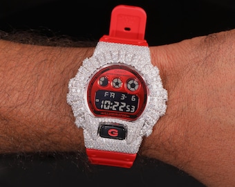 May include: A red and silver Casio G-Shock watch with a digital display showing the time as 10:22:53. The watch features a red band and a bezel encrusted with sparkling stones. The watch is on a wrist.