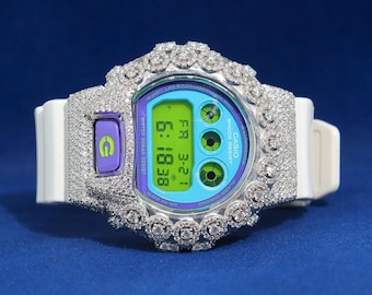 May include: A white Casio digital watch with a diamond-encrusted bezel and accents. The watch face displays the time 6:18:38 and the date. The watch has a white band and a purple button.