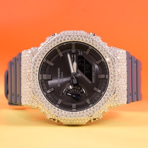 以下が含まれることがあります： ダークグレーの文字盤とグレーのバンドが付いたG-Shockの腕時計。ベゼルにはキラキラ光る石がちりばめられています。文字盤には時刻と日付が表示されています。