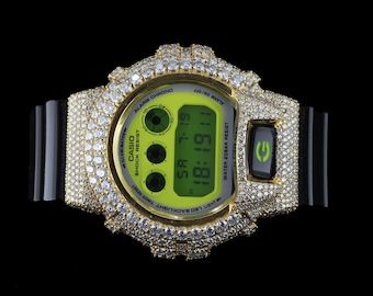 May include: A gold-toned Casio digital watch with a black band, encrusted with sparkling stones. The watch face is lime green, displaying the time and date. The watch has the text "CASIO SHOCK RESIST" on the face.