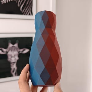 Könnte beinhalten: Eine geometrische Vase mit facettiertem Design, die einen Farbverlauf aus Blau- und Rottönen aufweist. Die Vase wird in einer Hand gehalten und zeigt ihre einzigartige Form und Farbgebung. Geeignet für die Inneneinrichtung.
