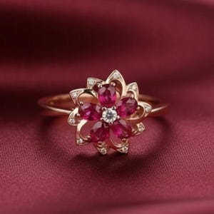 Puede incluir: Un anillo dorado con un diseño floral. Los pétalos de la flor están formados por piedras preciosas ovaladas de color rojo intenso, con una pequeña piedra preciosa clara y redonda en el centro. Pequeñas piedras preciosas claras acentúan los bordes exteriores de la flor.